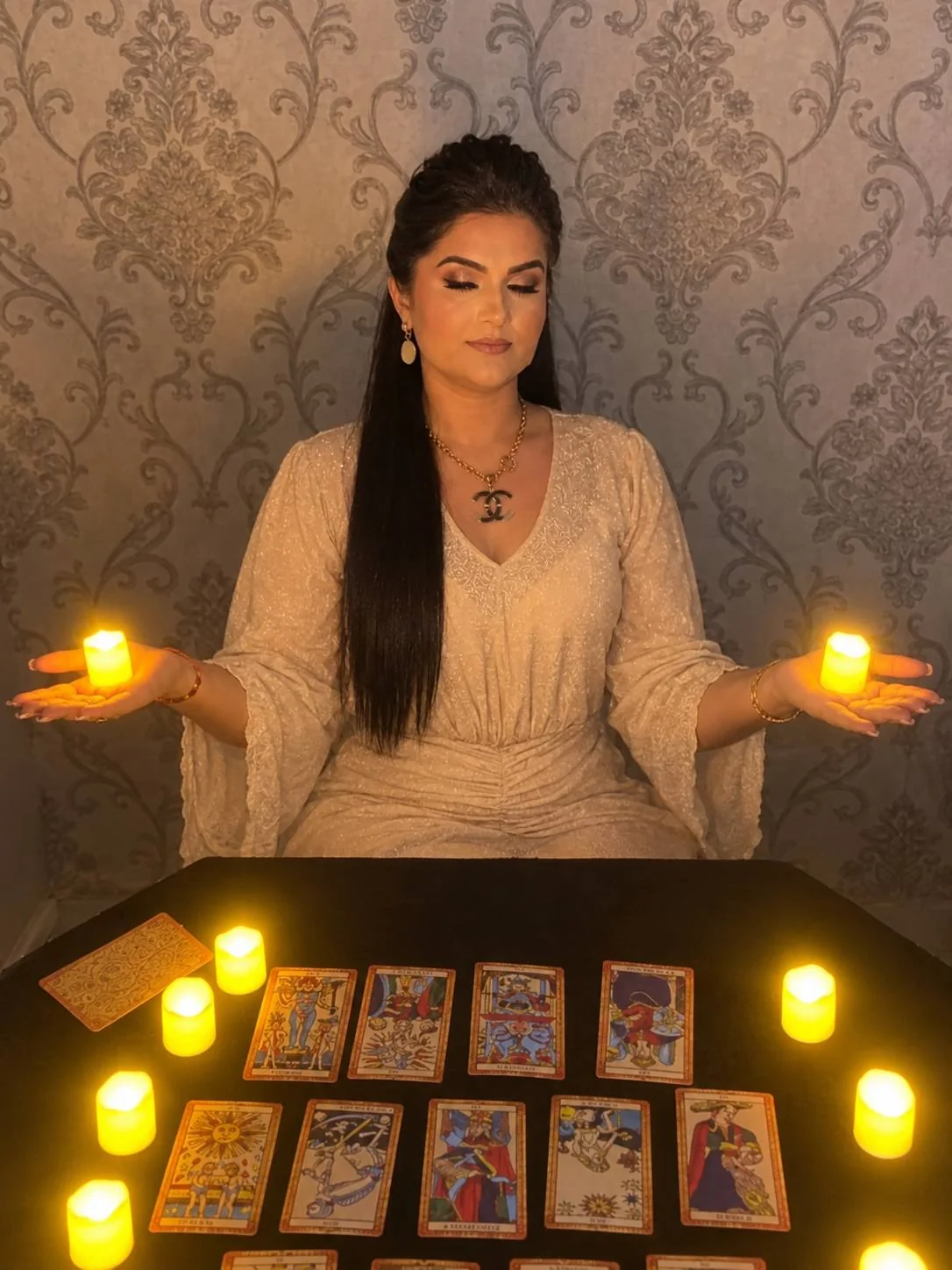 Sensitiva Cristina sentada à mesa de atendimento, de olhos fechados, com uma vela acesa em cada palma e cartas abertas à frente